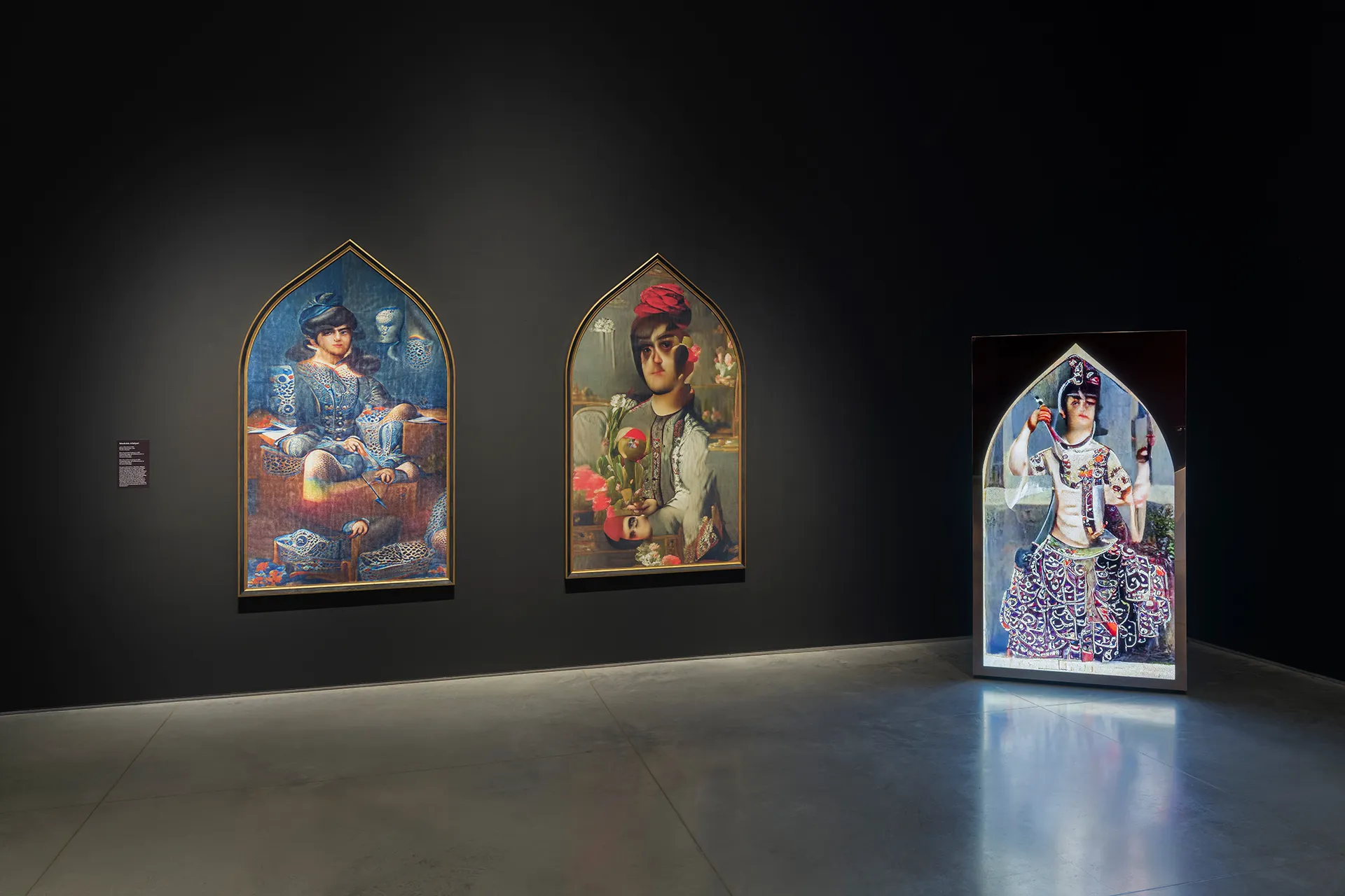 Gallery interior with black walls and a gray floor. On the left, two framed portraits hang side by side on the wall. On the right, a monitor standing on the floor displays a digital portrait.
