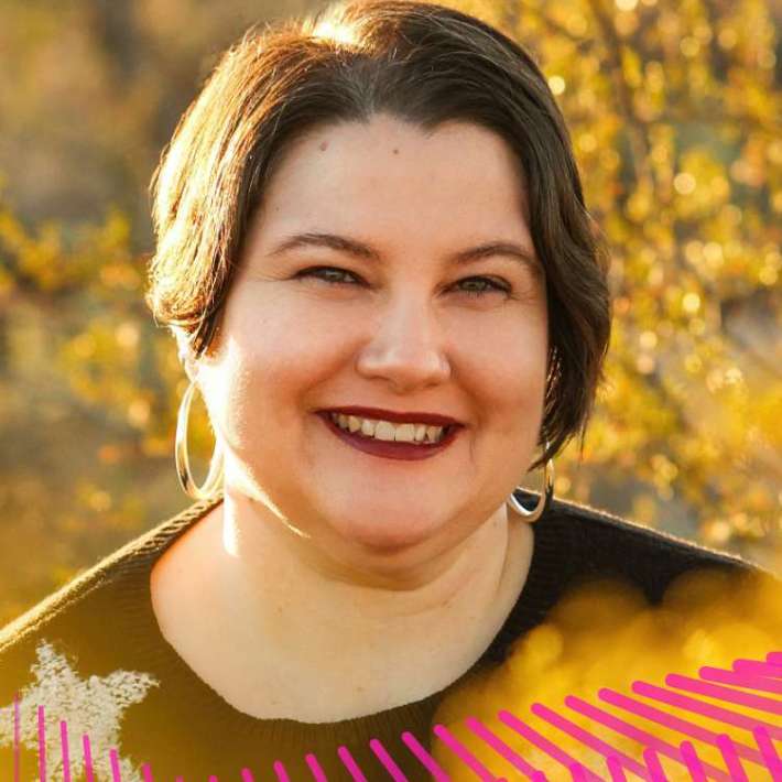 A light-skinned Mestizx with dark hair, silver hoop earrings, burgundy lipstick, and a black sweater with a white star. They sit in their scooter smiling and surrounded by golden creosote bushes.