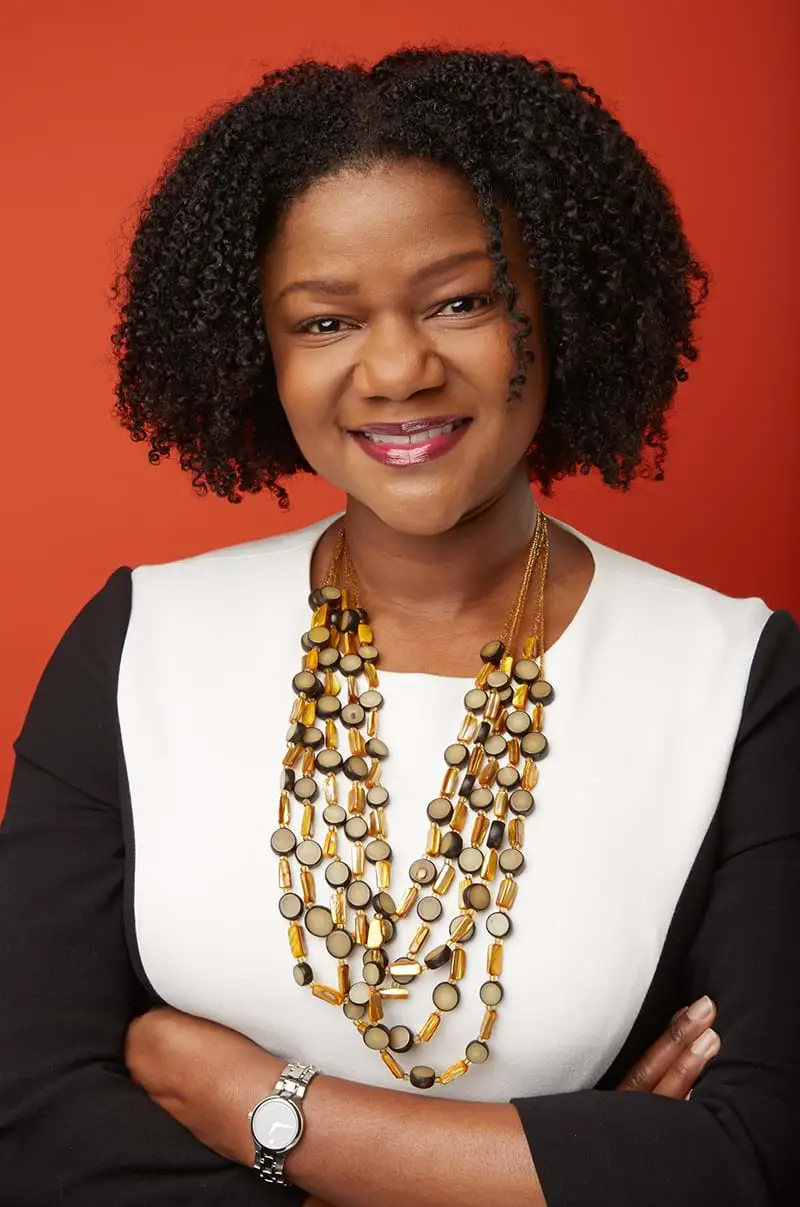 Diane Samuels in a black and white blouse and layered necklace against a red backdrop.