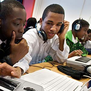 Two Black youth work on computers and music equipment. One wears headphones and taps their hand on a piano keyboard.
