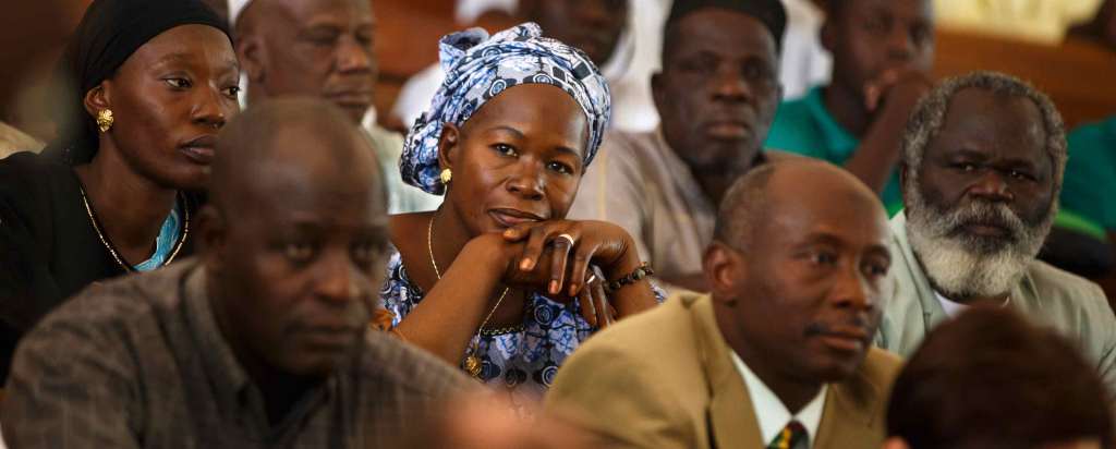 Black adult community members attend a class. The camera focuses on a person with a blue and grey patterned head wrap and matching shirt with gold jewelry.