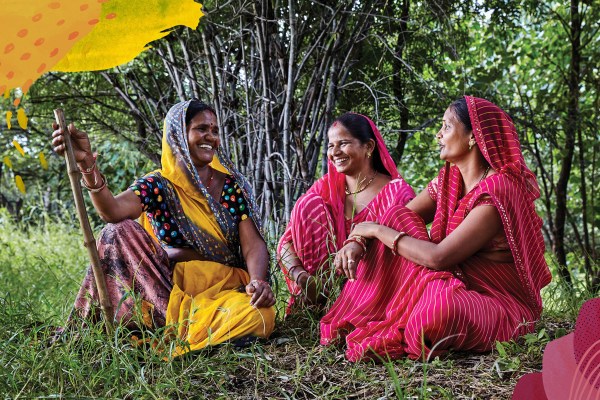 Three Indian women in mostly red garb sitting together under trees.
